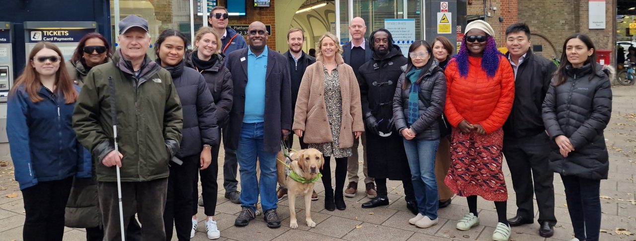 Group shot of Wandsworth Council’s Road Safety and Engineering Team with Sight Loss Council (SLC) members and Thomas Pocklington Trust staff. A selection of the team who have just been guided are wearing simulation glasses (sim specs). SLC member Harry is holding a navigation cane, and SLC member Haren is pictured with a guide dog. They are stood in front of Wandsworth Town train station.