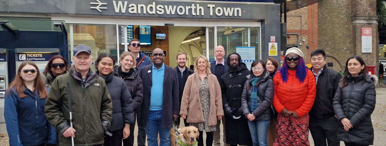 Group shot of Wandsworth Council’s Road Safety and Engineering Team with Sight Loss Council (SLC) members and Thomas Pocklington Trust staff. A selection of the team who have just been guided are wearing simulation glasses (sim specs). SLC member Harry is holding a navigation cane, and SLC member Haren is pictured with a guide dog. They are stood in front of Wandsworth Town train station.