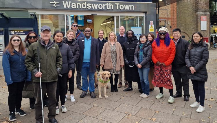 Group shot of Wandsworth Council’s Road Safety and Engineering Team with Sight Loss Council (SLC) members and Thomas Pocklington Trust staff. A selection of the team who have just been guided are wearing simulation glasses (sim specs). SLC member Harry is holding a navigation cane, and SLC member Haren is pictured with a guide dog. They are stood in front of Wandsworth Town train station.