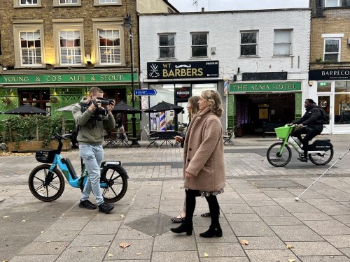 Camera man walking backwards next to an e-bike parked on the pavement with a film camera. He is filming a Thomas Pocklington Trust Sight Loss Council staff member walking down the street with a reporter who is wearing sim specs. The photo shows their side view walking down the pedestrianised area. Shops are in the background, and someone riding an e-bike.