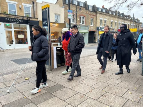 Members of Wandsworth Council’s Road Safety and Engineering Team walking down a street in Wandsworth Town in pairs. One is wearing simulation glasses and the other in the pair is guiding them. They are walking on a wide pavement in a pedestrianised area. Shops and a sign are in the background.