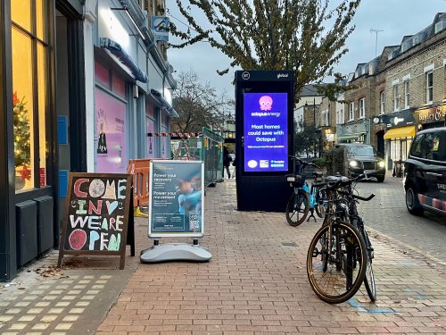 A pavement showing street obstacles including two advertising boards to the left, a parked bike to the right, a bus stop and a tree in the background. Roadworks can also be seen to the left side of the pavement in the background.