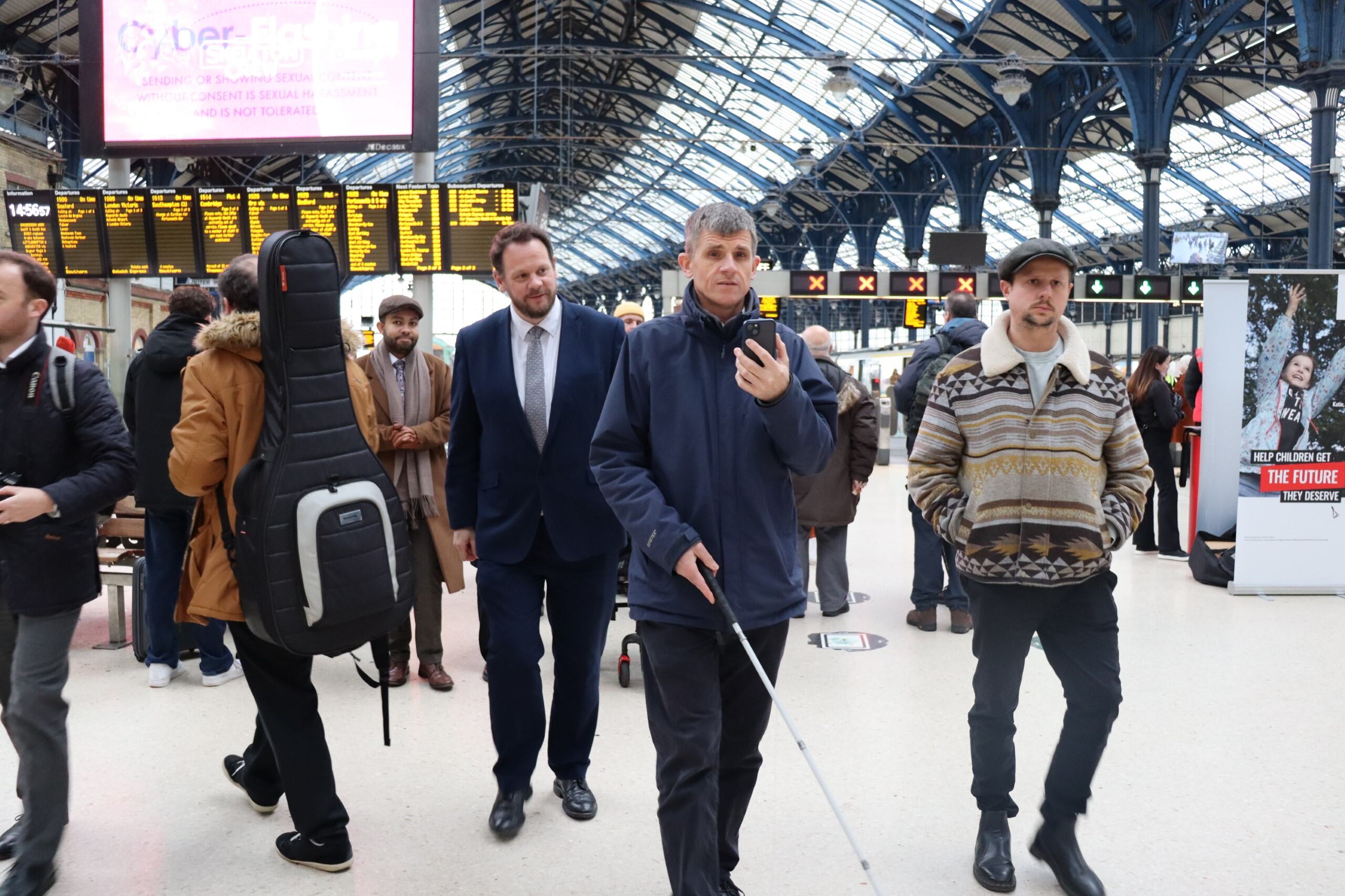 Dave Smith demonstrating the AIRA app inside a station to the Local Transport Minister in Brighton. He is walking holding his smartphone which he uses to speak to an AIRA operator. Transport Minister Simon Lightfoot walks behind him. In the background are crowds of people and passengers, and the stations electronic information boards.