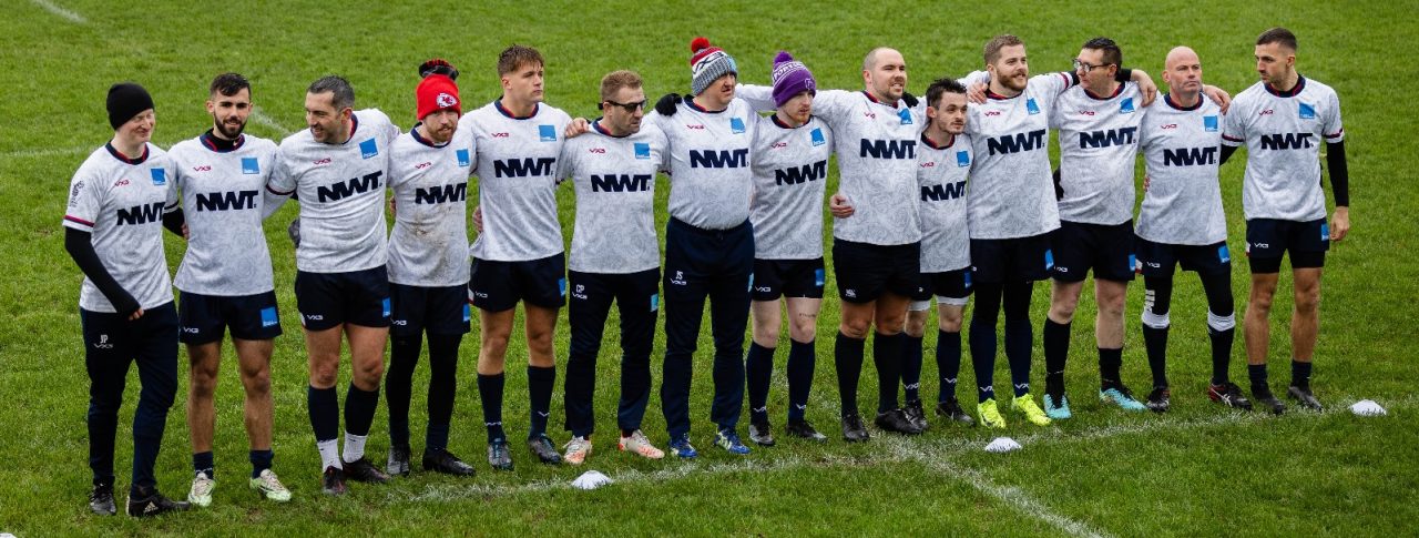 The England Vi rugby team standing in a line on a rugby pitch. The team are dressed in white rugby t-shirts and black shorts, with some wearing headbands and hats.