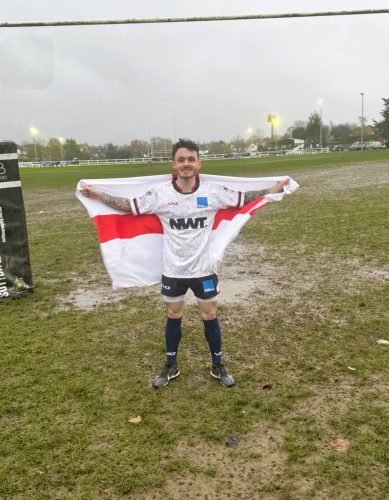 David Parfett waving the England flag, standing on a rugby pitch. David is wearing his VI rugby kit consisting of a white t shirt with a dark collar and black shorts. 