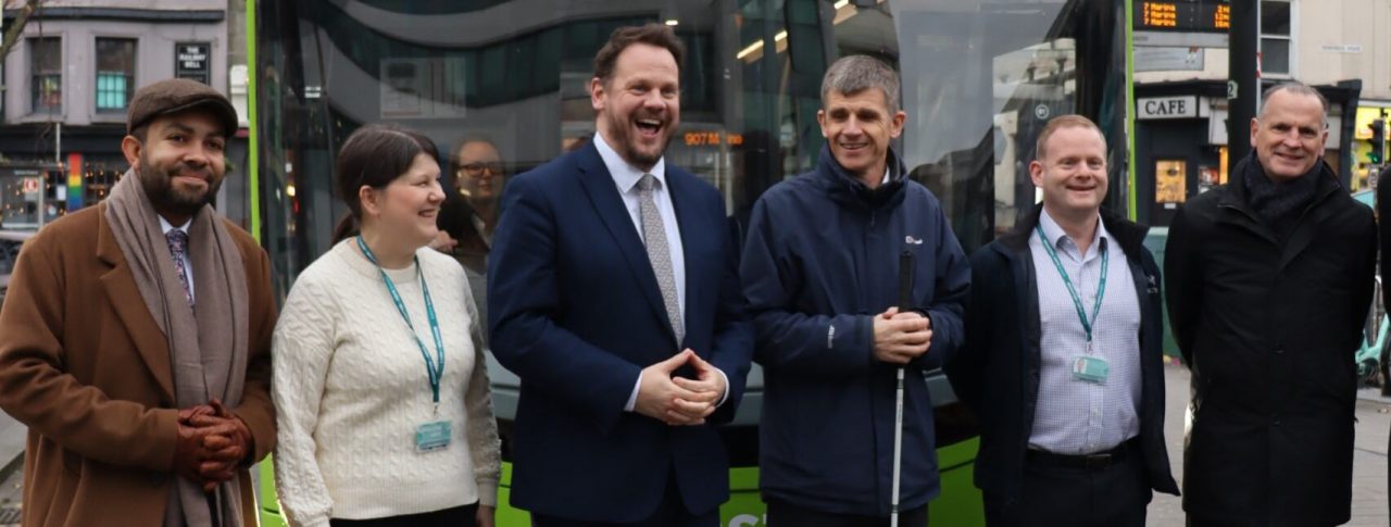 Dave stood with Transport Minister Simon Lightwood, Victoria Garcia, Ed Wills and another outside of Coaster bus at a bus stop. They are stood infront of the bus and smiling a the camera. The green bus says 'Accessible Travel' and the words 'Stephen Barnwell' at the top.