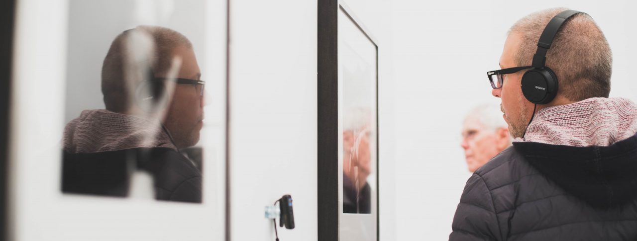 A man wearing headphones and glasses, standing in an art gallery. They are viewing a piece of art displayed on a wall, reflected in a nearby mirror.