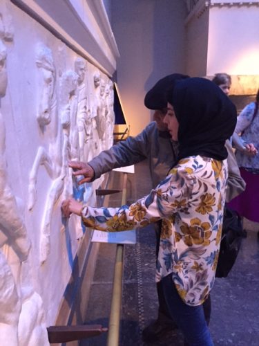 A woman wearing a floral shirt and hijab interacting with a tactile display on a wall featuring raised artwork of figures and animals. 