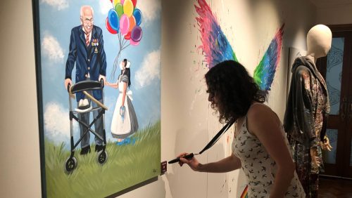 A woman standing opposite a painting is holding a long cane in her left hand and has a lanyard with a fob pressed against a box to the bottom of right of the artwork. The painting depicts an elderly man with a walking stick dressed in a suit and adorned with medals. holding hands with a girl in a maids uniform who is holding colourful balloons.