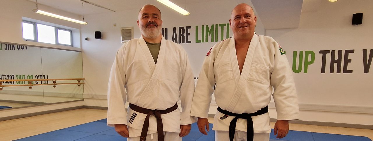 Two men wearing Judo outfits standing on a blue mat in a sports hall.