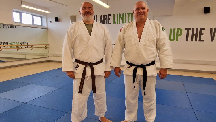 Two men wearing Judo outfits standing on a blue mat in a sports hall.