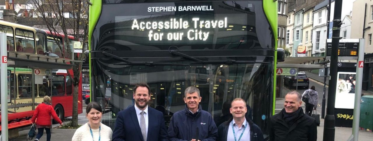 Dave stood with Transport Minister Simon Lightwood, Victoria Garcia, Ed Wills and another outside of Coaster bus at a bus stop. They are stood infront of the bus and smiling a the camera. The green bus says 'Accessible Travel' and the words 'Stephen Barnwell' at the top.