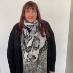 Elaine Dampier headshot. Elaine is standing against a white wall, has long dark hair, and is wearing a black cardigan and black and white scarf.