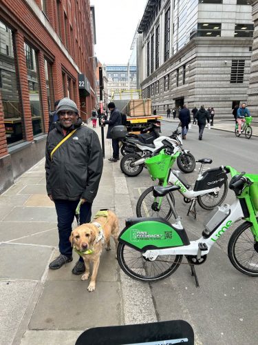 SW Volunteer Haren standing next three Lime bikes with one parked onto the curb of the road with his guide dog.