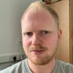 James Langley headshot. James has fair hair and a beard and is wearing a grey t-shirt. He is facing the camera, smiling.