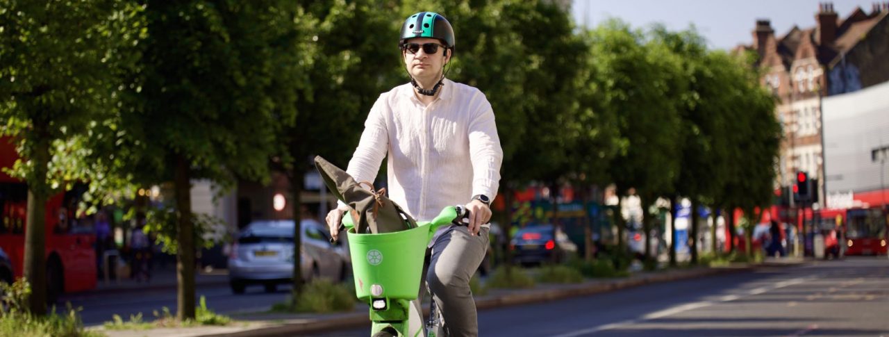 Man riding a green Lime bike on a sunny street with trees and buildings in the background. The man is dressed in a pink shirt and brown trousers and is wearing a green helmet and black sunglasses.