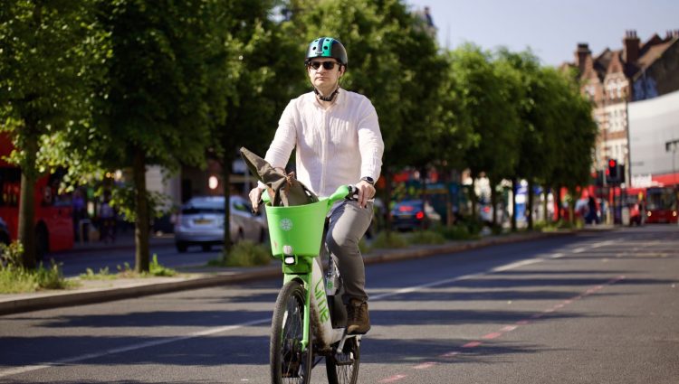 Man riding a green Lime bike on a sunny street with trees and buildings in the background. The man is dressed in a pink shirt and brown trousers and is wearing a green helmet and black sunglasses.