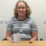 Linn Davies headshot. Linn is wearing a white and black striped t-shirt and sitting at a table while smiling at the camera.