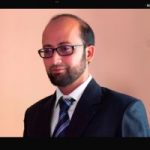 Mohammed Salman Shaikh headshot. Mohammed has short black hair and is wearing glasses, a black suit and white shirt with a navy tie. He is smiling at the camera.