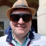 Paul Goddard. Paul is wearing a straw hat, sunglasses and a white and navy coat while smiling at the camera.