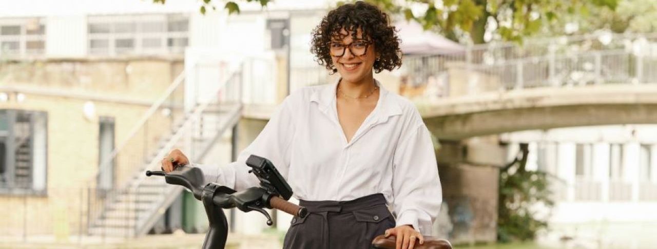 Woman dressed in smart attire posing with a dark green Forest bike on a leafy street. She is dressed in a white shirt and long black trousers and has short black hair.