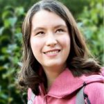 Ellie Wallwork headshot. Ellie has brown hair and is wearing a pink hoodie while smiling at the camera with trees in the background.