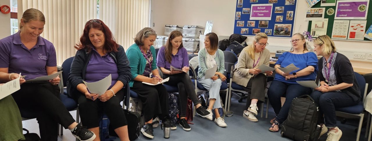 Eight women, all members staff members of Sirona Healthcare, are reading from paper handouts and talking amongst themselves. All of them are seated are are in a classroom style room.