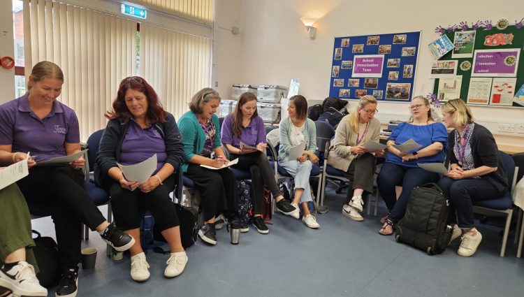 Eight women, all members staff members of Sirona Healthcare, are reading from paper handouts and talking amongst themselves. All of them are seated are are in a classroom style room.