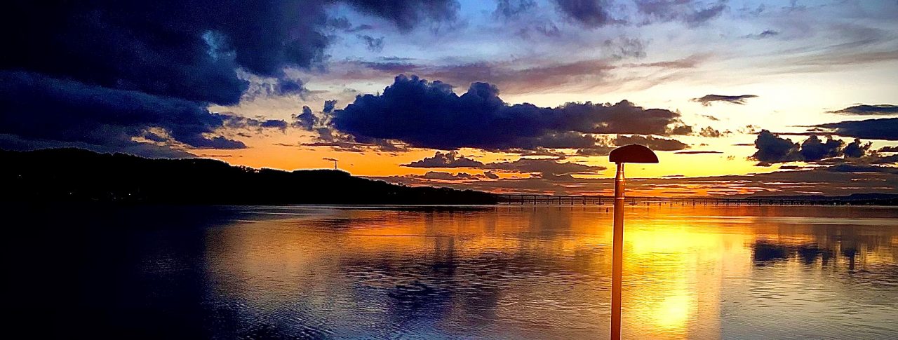 A striking painting of a sunset over a calm lake. The sky is a dramatic mix of deep blue clouds and warm orange, pink and yellow hues reflecting on the water. A single lamppost stands near the water’s edge, silhouetted against the sky.