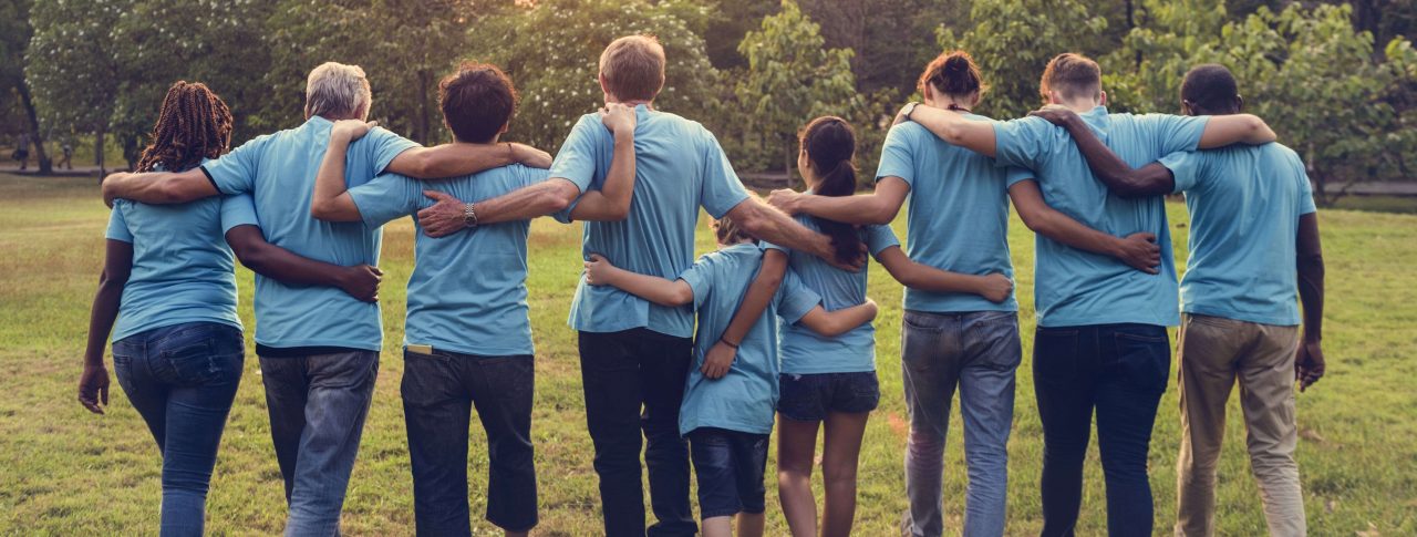 Group of volunteers with their arms around each other, facing away from the camera. They are outside and are all wearing light blue t-shirts.